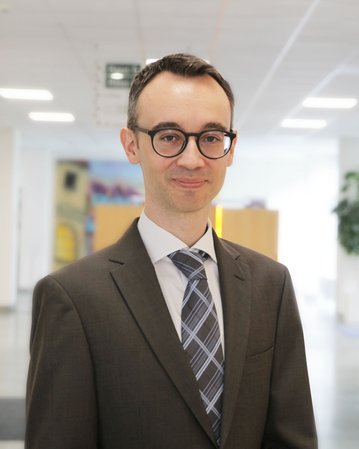 Ein Mann in einem Anzug mit Brille steht in einem modernen Büro. Er hat kurzes, dunkles Haar und trägt eine gestreifte Krawatte.