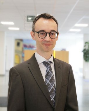 Ein Mann in einem Anzug mit Brille steht in einem modernen Büro. Er hat kurzes, dunkles Haar und trägt eine gestreifte Krawatte.