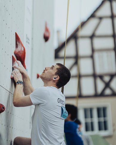 Ein Kletterer in einem weißen T-Shirt greift nach roten Griffen an einer Kletterwand, während ein weiterer Kletterer im Hintergrund sichtbar ist.