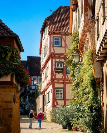 Paar, das auf einer gepflasterten Straße in einer malerischen Altstadt spaziert, umgeben von historischen Fachwerkhäusern und Pflanzen.