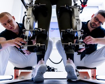 Zwei Techniker arbeiten an einem Exoskelett, das an den Beinen einer Person befestigt ist, während sie Anpassungen vornehmen.