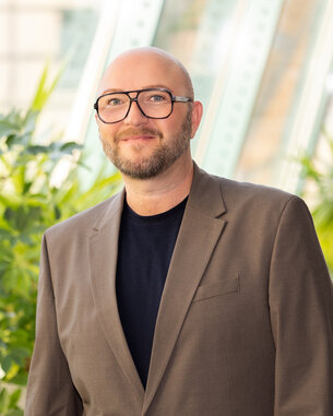 Mann mit Brille und Anzug steht in einem modernen Raum mit Pflanzen im Hintergrund.