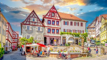 Fachwerkhäuser und ein Brunnen in einem malerischen Platz, umgeben von Cafés und Fahrrädern, unter einem bunten Himmel.