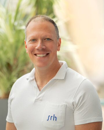 Ein Mann mit kurzem Haar trägt ein weißes Polo-Shirt mit dem Logo "srh" und lächelt vor einer grünen Pflanzenkulisse.