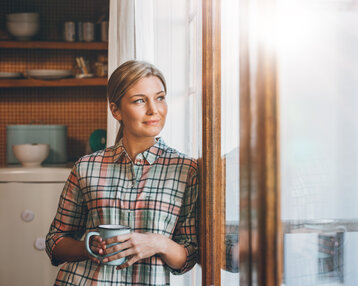 Eine Frau mit einem Kaffeebecher lehnt an einem Fenster und schaut nachdenklich nach draußen. Sie trägt ein kariertes Hemd.