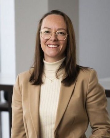 Eine lächelnde Frau mit langen, braunen Haaren trägt eine beige Blazerjacke und eine helle Bluse. Sie steht in einem modernen Büro.