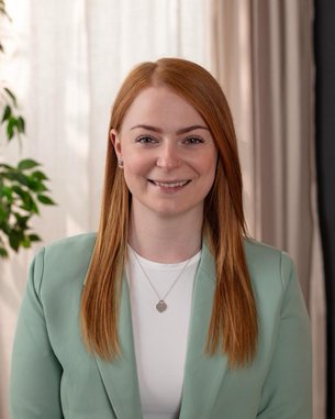 Eine lächelnde Frau mit langen, roten Haaren trägt einen hellgrünen Blazer und steht vor einem hellen Hintergrund mit Pflanzen.