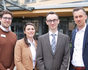 Vier Personen stehen zusammen vor einem Gebäude. Sie tragen formelle Kleidung und lächeln in die Kamera.