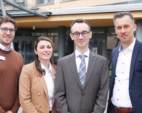 Vier Personen stehen zusammen vor einem Gebäude. Sie tragen formelle Kleidung und lächeln in die Kamera.