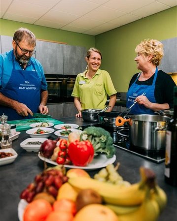 Drei Personen in einer Küche bereiten gemeinsam eine Mahlzeit vor. Frisches Gemüse und Obst sind auf dem Tisch angerichtet.