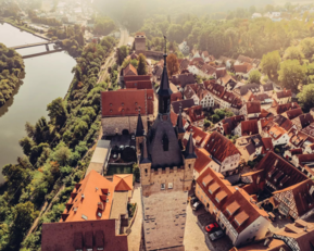 Luftaufnahme einer Stadt mit einem markanten Kirchturm, umgeben von roten Dächern und grünen Bäumen, neben einem Fluss.