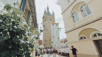 Eine Kletterwand steht zwischen historischen Gebäuden, während Menschen daran klettern und sich im Freien aufhalten. Im Hintergrund ist ein Turm sichtbar.