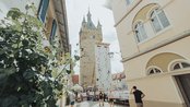 Eine Kletterwand steht zwischen historischen Gebäuden, während Menschen daran klettern und sich im Freien aufhalten. Im Hintergrund ist ein Turm sichtbar.
