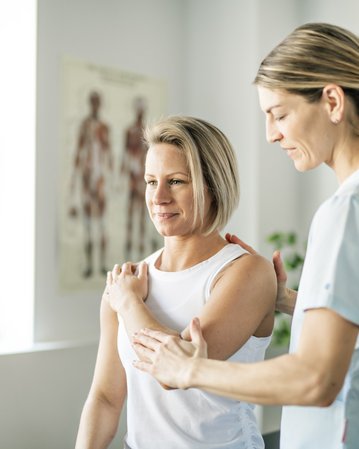 Eine Physiotherapeutin führt eine Behandlung bei einer Patientin durch, die auf einer Bank sitzt. Die Therapeutin berührt sanft die Schulter der Patientin, während sie Anweisungen gibt. Im Hintergrund sind anatomische Modelle sichtbar.