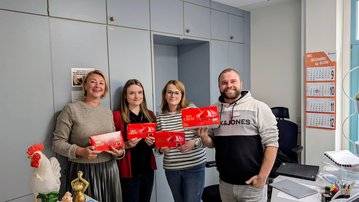 Robert Koch Krankenhaus Stuttgart zu Besuch Vier Personen stehen in einem Büro und halten rote Verpackungen in den Händen. Im Hintergrund sind Büromöbel und ein Kalender sichtbar.