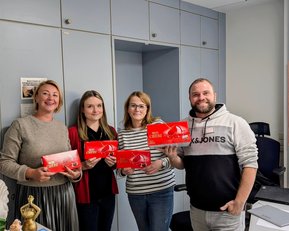 Robert Koch Krankenhaus Stuttgart zu Besuch Vier Personen stehen in einem Büro und halten rote Verpackungen in den Händen. Im Hintergrund sind Büromöbel und ein Kalender sichtbar.