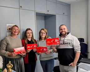 Vier Personen stehen in einem Büro und halten rote Verpackungen in den Händen. Im Hintergrund sind Büromöbel und ein Kalender sichtbar.