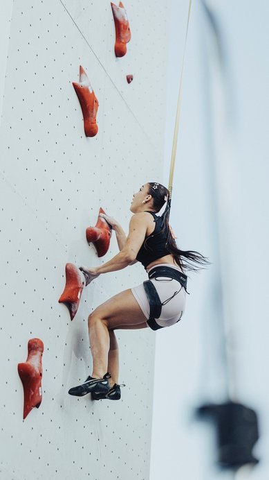 Eine Kletterin erklimmt eine vertikale Kletterwand mit roten Griffen, gesichert durch ein Seil. Sie trägt sportliche Kleidung und Kletterschuhe.