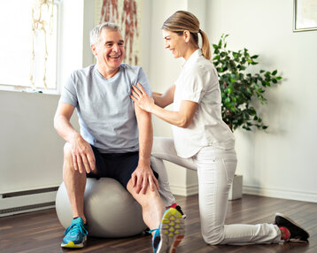 Ein älterer Mann sitzt auf einem Gymnastikball, während eine Physiotherapeutin ihm freundlich hilft und ihn an der Schulter berührt.