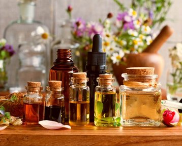 Eine Anordnung von verschiedenen Fläschchen mit ätherischen Ölen und Kräutern auf einem Holztisch, umgeben von frischen Blumen und einem Mörser. Die Fläschchen haben unterschiedliche Größen und Formen, einige sind mit Korken verschlossen.