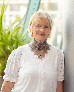 Eine Person mit blonden Haaren und Brille steht neben einer Säule, umgeben von Pflanzen. Sie trägt ein weißes Oberteil und hat sichtbare Tattoos.