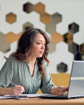 Eine Frau sitzt an einem Tisch, arbeitet am Laptop und macht Notizen. Im Hintergrund sind hexagonale Wandpaneele sichtbar.