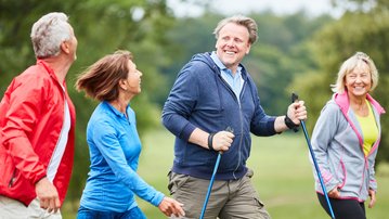 Eine Gruppe von vier Personen geht gemeinsam auf einem Weg. Sie tragen Sportkleidung und nutzen Nordic-Walking-Stöcke. Die Atmosphäre wirkt aktiv und gesellig, während sie sich unterhalten und lächeln.