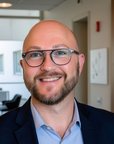 Ein Mann mit Brille und Bart trägt einen dunklen Anzug und ein hellblaues Hemd. Er steht in einem modernen Büro mit hellen Wänden und Möbeln im Hintergrund.