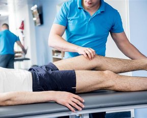 Ein Physiotherapeut behandelt das Bein eines Patienten auf einer Liege, während ein weiterer Patient im Hintergrund an einer Übung arbeitet.