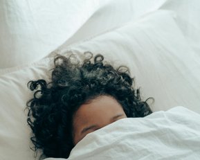 Ein Kind mit lockigem Haar liegt im Bett und ist teilweise mit einer Decke zugedeckt.