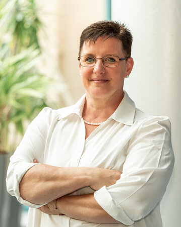 Eine Person mit kurzen Haaren und Brille steht mit verschränkten Armen vor einer Säule, umgeben von Pflanzen in einem modernen Raum.