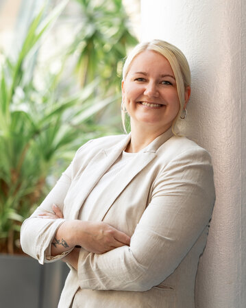 Eine lächelnde Frau mit blonden Haaren steht an einer Säule und hat die Arme verschränkt. Im Hintergrund sind Pflanzen sichtbar.