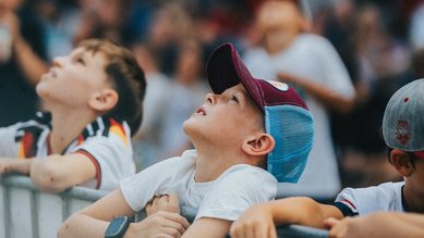 Kinder stehen an einem Zaun und schauen nach oben, während sie auf ein Event warten. Einige tragen Caps und T-Shirts.