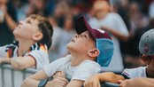 Kinder stehen an einem Zaun und schauen nach oben, während sie auf ein Event warten. Einige tragen Caps und T-Shirts.