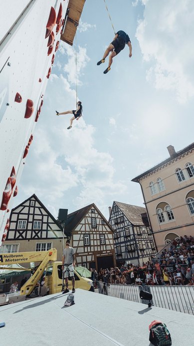 Zwei Sportler beim Klettern an einer Wand, einer schwingt in der Luft, während Zuschauer auf einer Tribüne zuschauen.