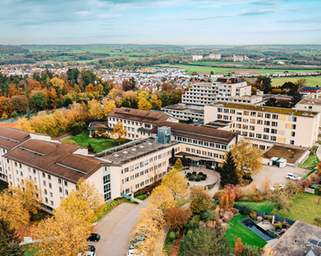 Ein weitläufiges Gebäudeensemble in einer herbstlichen Landschaft, umgeben von bunten Bäumen. Die Architektur umfasst mehrere miteinander verbundene Gebäude, die in einem ruhigen, ländlichen Gebiet liegen.