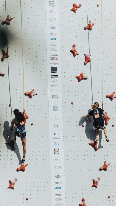 Zwei Kletterer in Sportkleidung erklimmen eine Kletterwand mit roten Griffen. Beide tragen Sicherheitsgurte und sind in einem Wettkampf.