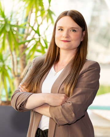 Eine Frau mit langen Haaren steht mit verschränkten Armen vor einer Fensterfront, umgeben von Pflanzen. Sie trägt einen blauen Blazer und ein weißes T-Shirt.