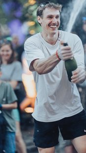 Ein Mann in einem weißen T-Shirt öffnet eine Champagnerflasche, während eine Gruppe von Menschen im Hintergrund zuschaut und lächelt.