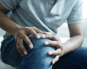 Eine Person sitzt auf einem Bett und hält sich mit beiden Händen das rechte Knie, möglicherweise aufgrund von Schmerzen oder Unbehagen.