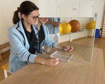 Eine Person sitzt an einem Tisch und betrachtet ihr Spiegelbild in einem großen Spiegel, während sie eine Hand auf den Tisch legt.