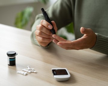 Eine Person hält einen Blutzuckermessgerät und eine Lanzette, während sie sich auf die Fingerkuppe vorbereitet. Neben ihnen liegen Teststreifen und ein Behälter.