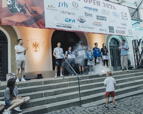 Eine Gruppe von Personen steht auf einer Bühne bei einer Veranstaltung, während ein Kind im Vordergrund spielt. Ein Banner mit Sponsoren ist im Hintergrund sichtbar.