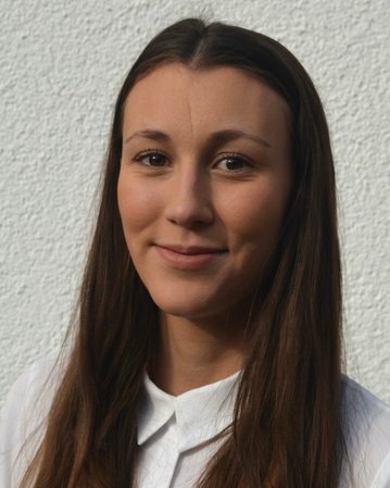 Eine junge Frau mit langen, braunen Haaren und einem weißen Hemd lächelt in die Kamera vor einer hellen Wand.