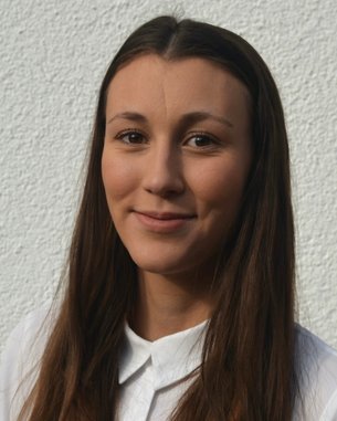 Eine junge Frau mit langen, braunen Haaren und einem weißen Hemd lächelt in die Kamera vor einer hellen Wand.