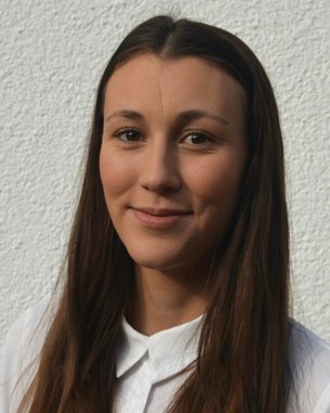 Eine junge Frau mit langen, braunen Haaren und einem weißen Hemd lächelt in die Kamera vor einer hellen Wand.