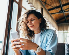 Eine Frau mit lockigem Haar sitzt in einem Café und schaut auf ihr Smartphone. Sie trägt ein lässiges Hemd und wirkt konzentriert.