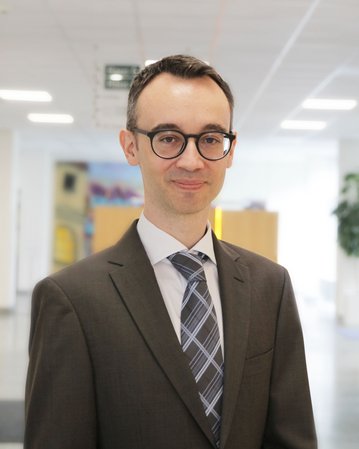 Ein Mann in einem Anzug mit Krawatte steht in einem modernen Büro. Er trägt eine Brille und lächelt leicht.