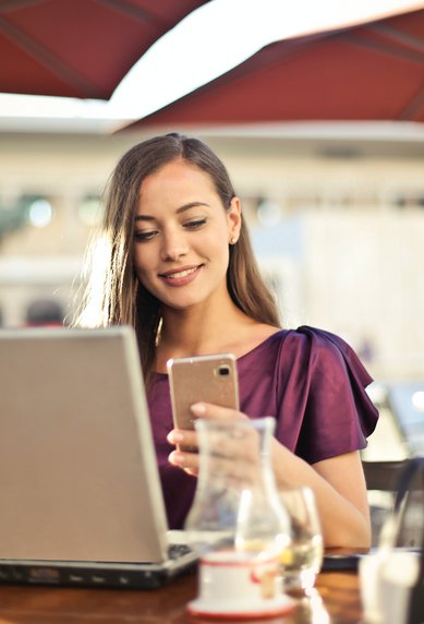 Eine Frau sitzt an einem Tisch in einem Café, lächelt und schaut auf ihr Smartphone, während ein Laptop vor ihr steht. Im Hintergrund sind Tische und Stühle sichtbar.