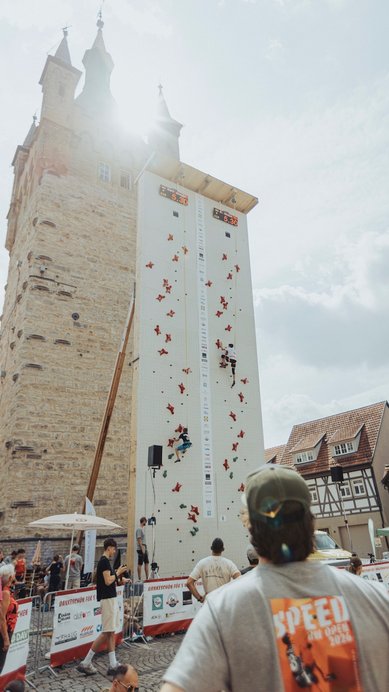 Kletterwettbewerb an einer hohen Wand mit Teilnehmern, die um die Wette klettern, während Zuschauer zuschauen. Historisches Gebäude im Hintergrund.